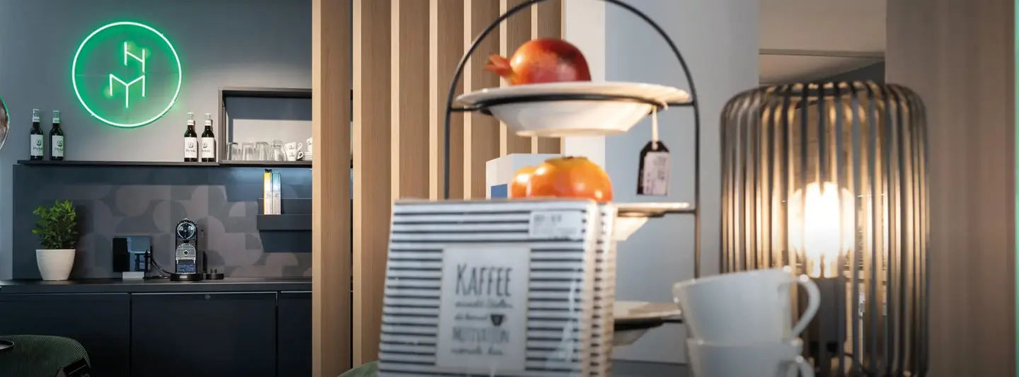 Dunkle Küchenzeile mit Regal, Kaffeemaschine und Flaschen, grünes Neonlogo an der Wand, Tisch und Lampe im Vordergrund